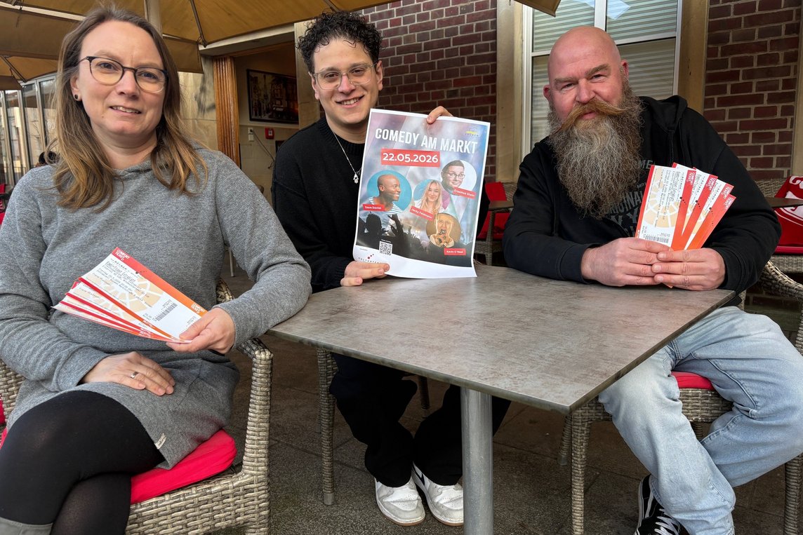 Das Bild zeigt drei Personen, die an einem Tisch sitzen und Flyer und ein kleines Plakat hochhalten.