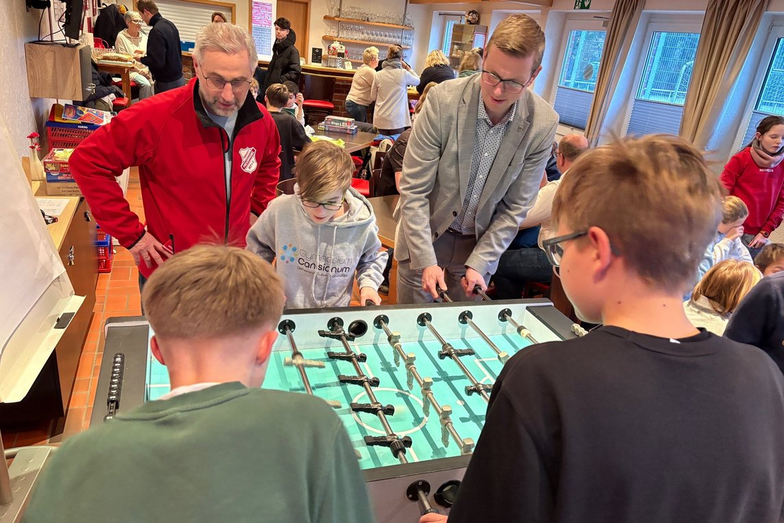 Das Bild zeigt insgesamt fünf Personen welche an einem Tischkicker spielen. 