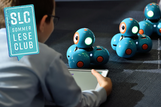 Ein Junge hält ein Tablet in der Hand und vor ihm stehen 3 Dash-Roboter.